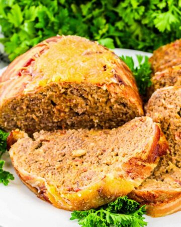 Close-up of crispy bacon-wrapped cheeseburger meatloaf topped with melted cheddar cheese, presented in a white baking dish with fresh parsley in the background.
