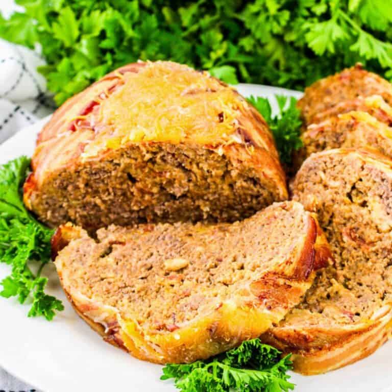 Close-up of crispy bacon-wrapped cheeseburger meatloaf topped with melted cheddar cheese, presented in a white baking dish with fresh parsley in the background.