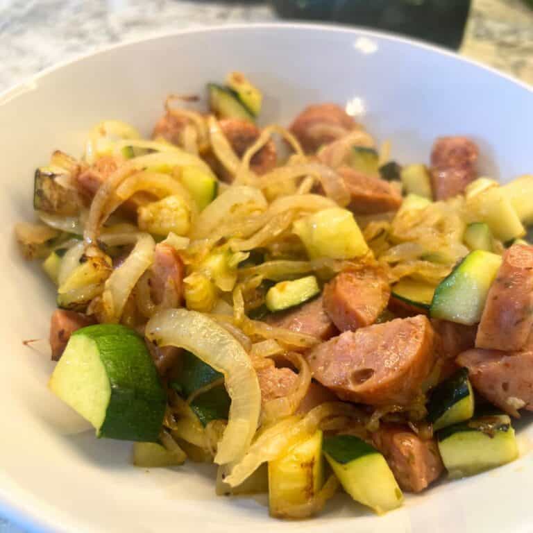 A white bowl filled with a freshly cooked sausage and zucchini skillet meal, showing browned sausage, tender zucchini, and caramelized onions.