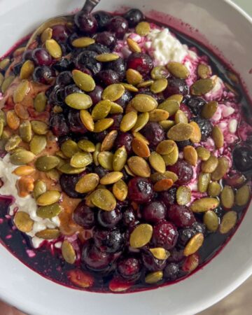 A white bowl filled with creamy cottage cheese, topped with warm, juicy blueberries, smooth almond butter, and crunchy roasted pumpkin seeds. A spoon rests inside the bowl, ready to enjoy this high-protein meal.