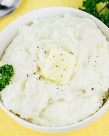 Bowl of cottage cheese mashed potatoes topped with a pat of butter and freshly ground pepper, on a yellow background.