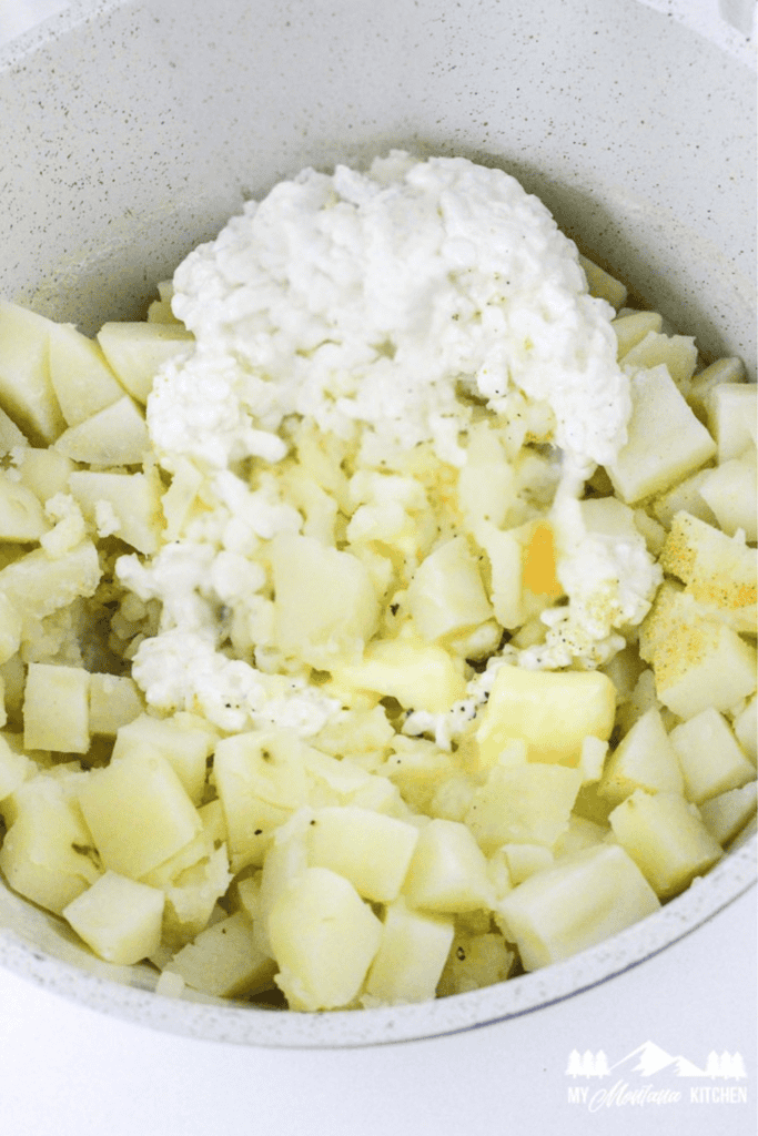 Cooked diced potatoes and a generous scoop of cottage cheese in a mixing pot, ready to be mashed.