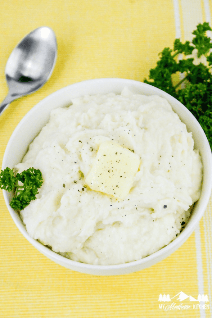 Bowl of finished mashed potatoes topped with melted butter and pepper, served with fresh parsley and a spoon on the side.