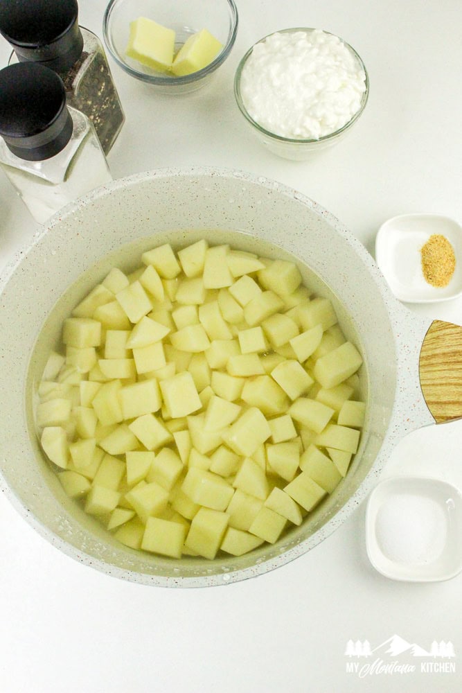 Diced raw potatoes submerged in water in a large pot, with cottage cheese, butter, and seasonings in the background.