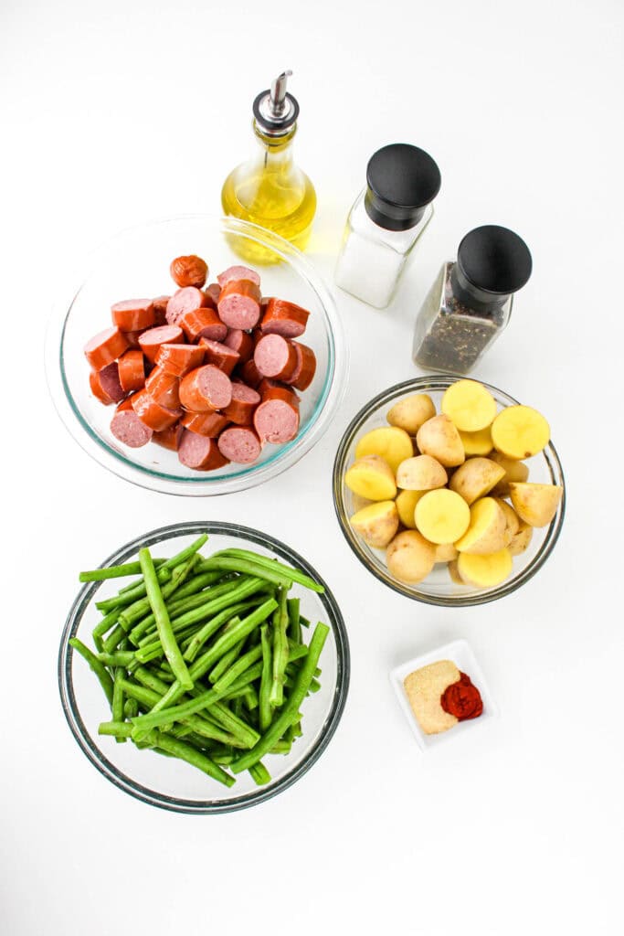 A top-down view of ingredients for a smoked sausage, green beans, and potatoes recipe, including sliced sausage, fresh green beans, halved baby potatoes, seasonings, olive oil, salt, and pepper.