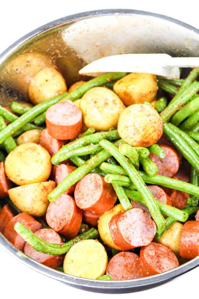 A large mixing bowl filled with uncooked baby potatoes, fresh green beans, and smoked sausage, ready to be seasoned.