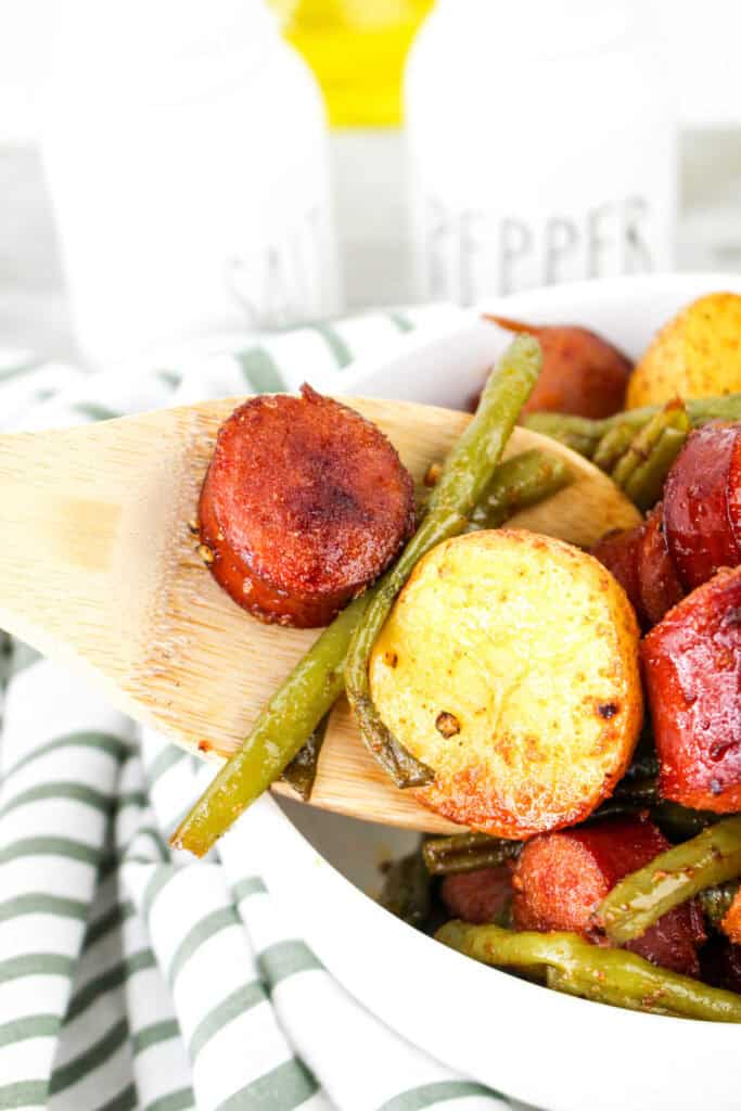 A close-up of a wooden spoon scooping up a serving of smoked sausage, golden potatoes, and green beans, highlighting the crispy texture and seasoning.