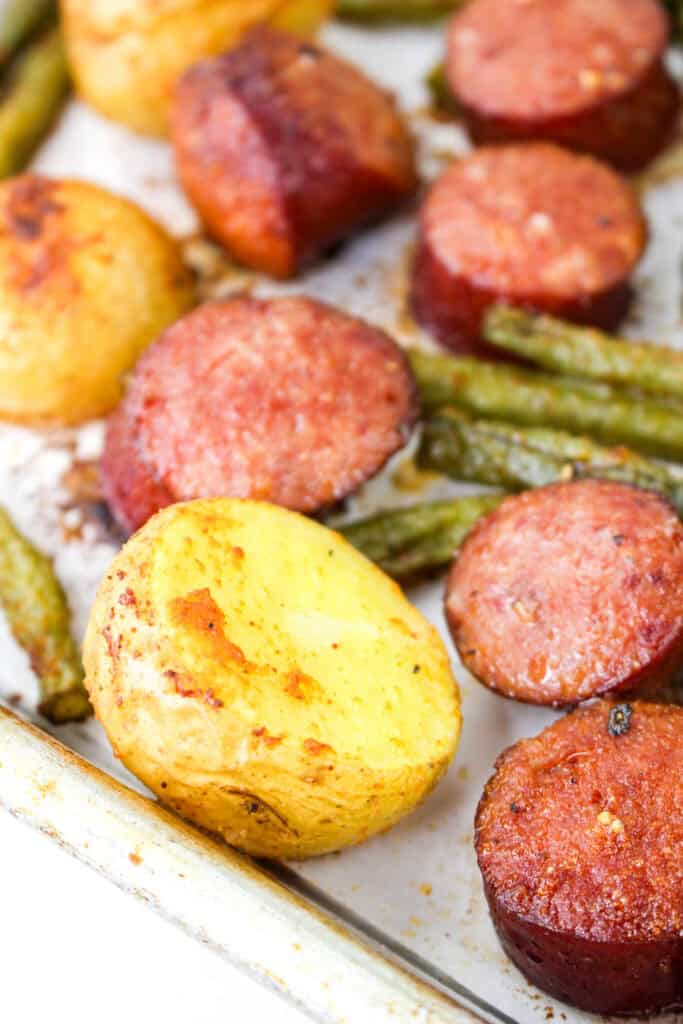 Close-up of crispy golden potato and browned smoked sausage on a baking sheet with roasted green beans.