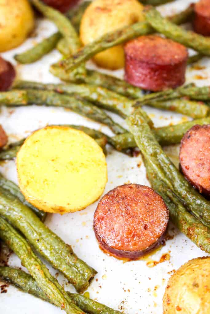 Close-up of roasted smoked sausage, golden potatoes, and green beans on a sheet pan, showing crispy textures and seasoning.