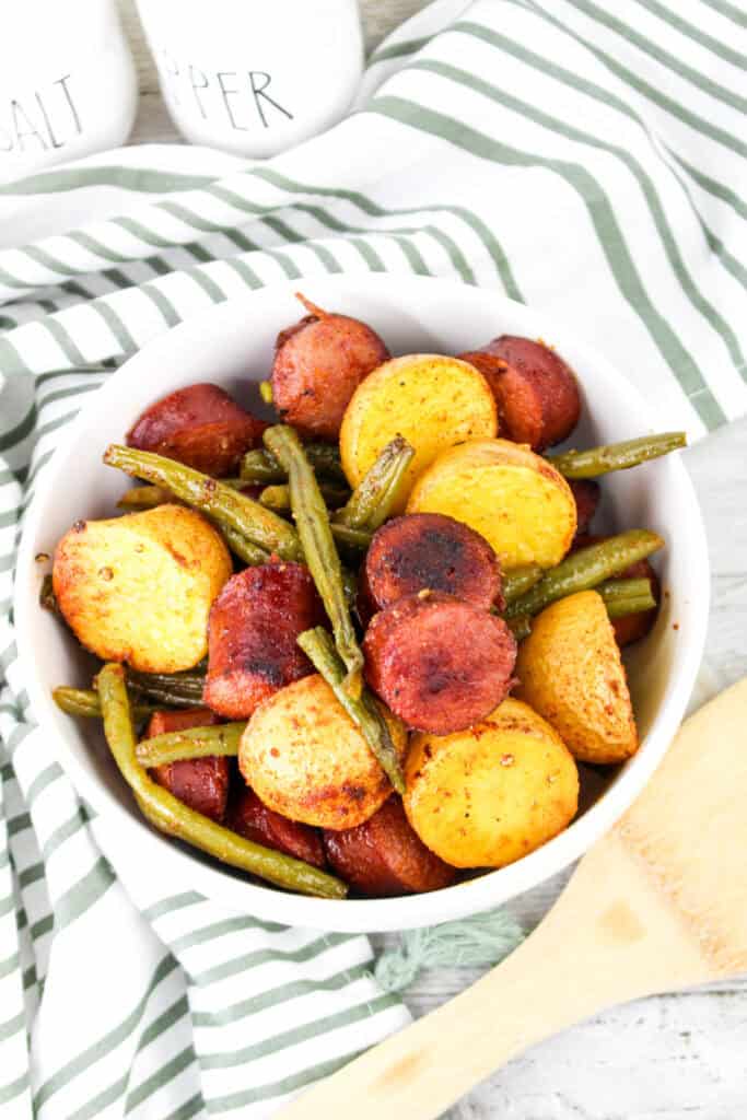 A beautifully arranged bowl of roasted sausage, golden baby potatoes, and green beans, with seasoning visible on the crispy edges of each ingredient.