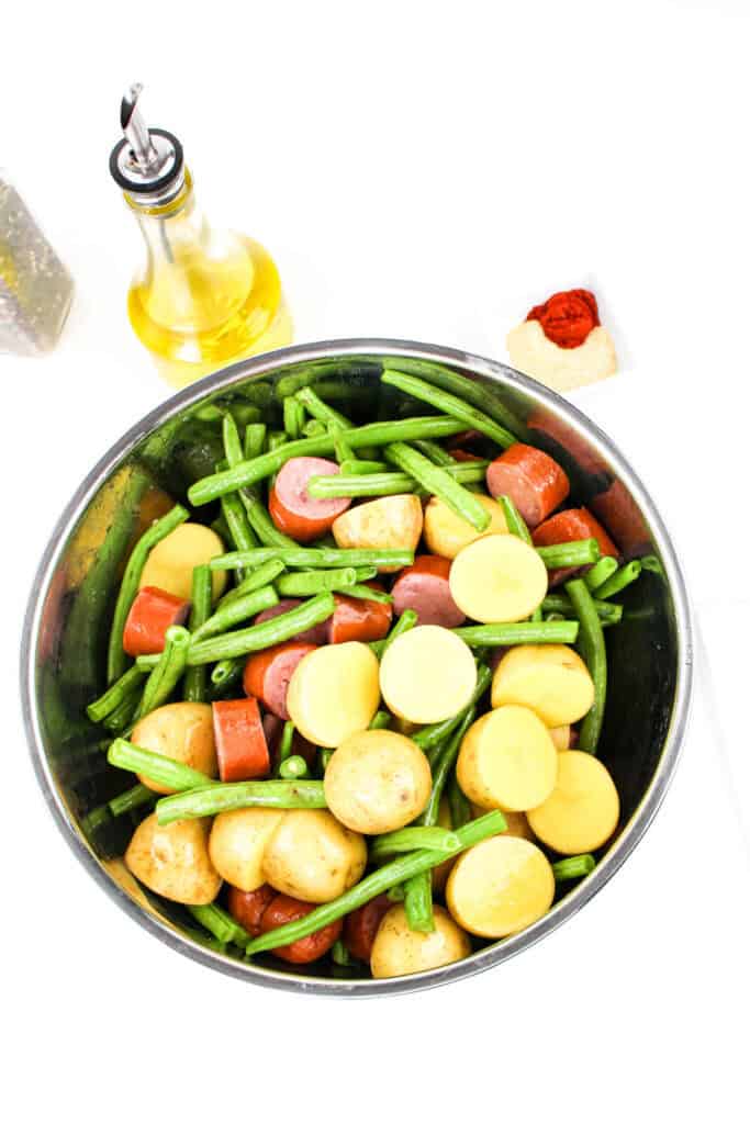 Overhead shot of a large mixing bowl filled with baby potatoes, fresh green beans, and smoked sausage, with seasonings and olive oil nearby.