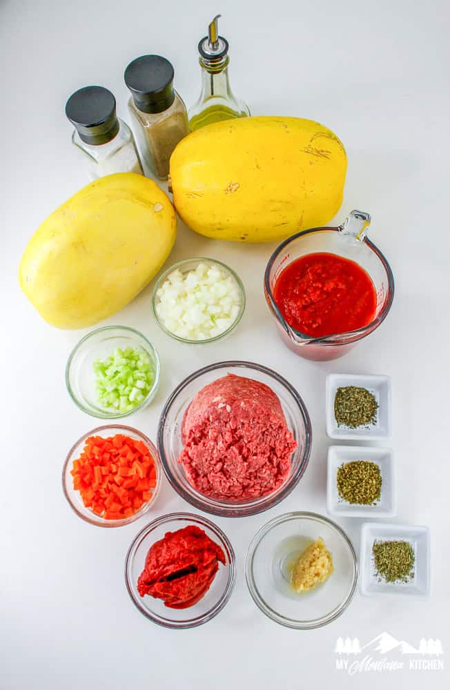 Flat lay of ingredients for spaghetti squash Bolognese including squash, ground beef, vegetables, tomato paste, herbs, and olive oil.