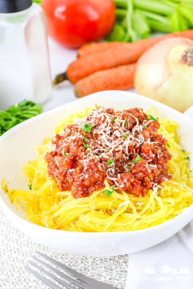 Side view of spaghetti squash topped with chunky Bolognese meat sauce and shredded cheese, surrounded by fresh veggies.