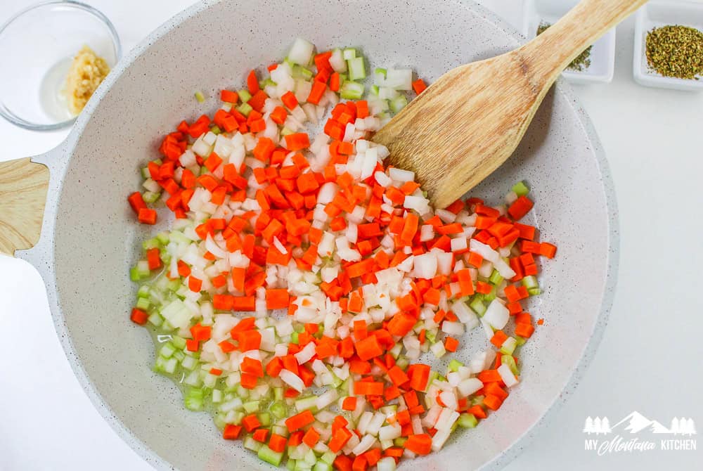 Carrots, celery, and onions being sautéed in a pan, stirred with a wooden spoon.