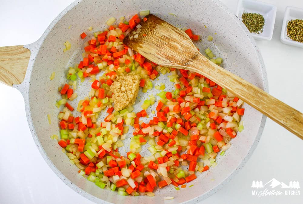 Minced garlic added to a sautéed vegetable mix of carrots, celery, and onion in a skillet with a wooden spoon.
