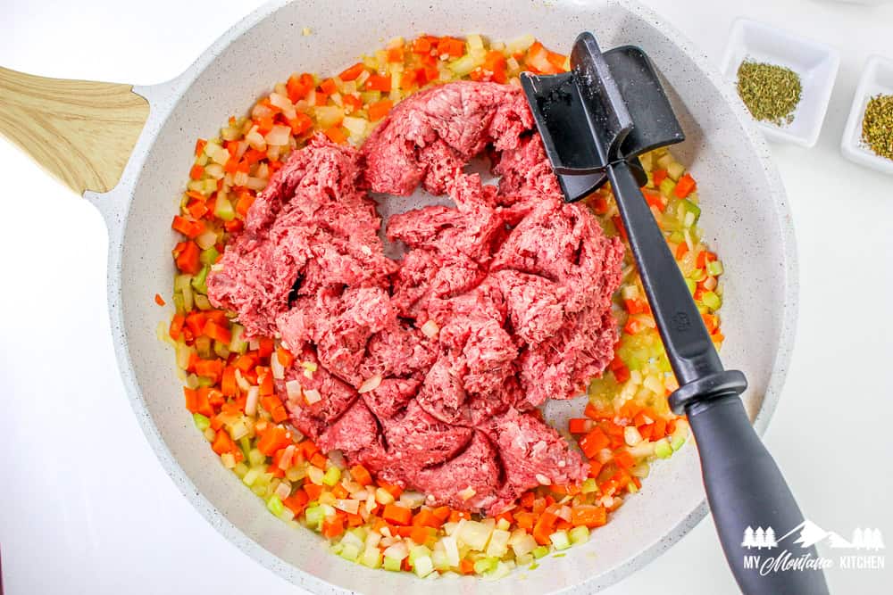 Raw ground beef added to a skillet filled with sautéed carrots, celery, and onions, ready to be browned.