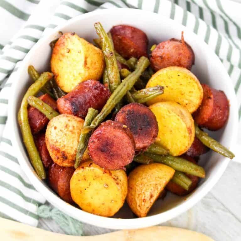 A close-up of a white bowl filled with roasted smoked sausage, crispy golden potatoes, and green beans, served on a striped green and white cloth.