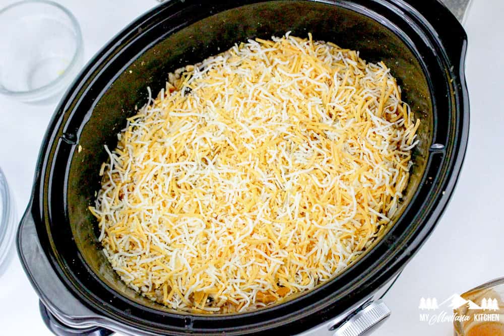 Crockpot filled with a layer of shredded cheese over enchilada casserole mixture.