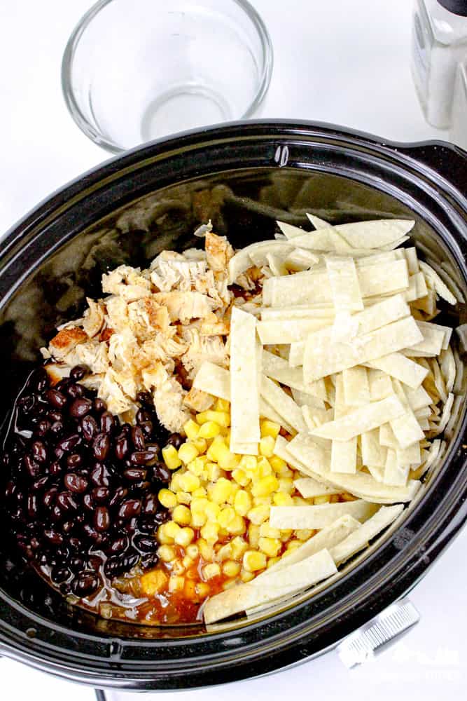 Layered crockpot ingredients including shredded chicken, black beans, corn, and tortilla strips before cooking.
