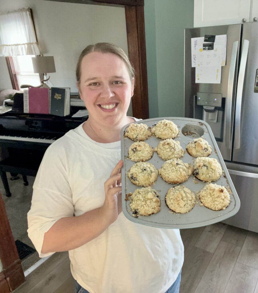 Woman holding a muffin tin of homemade sourdough blueberry muffins with crumb topping in a cozy kitchen