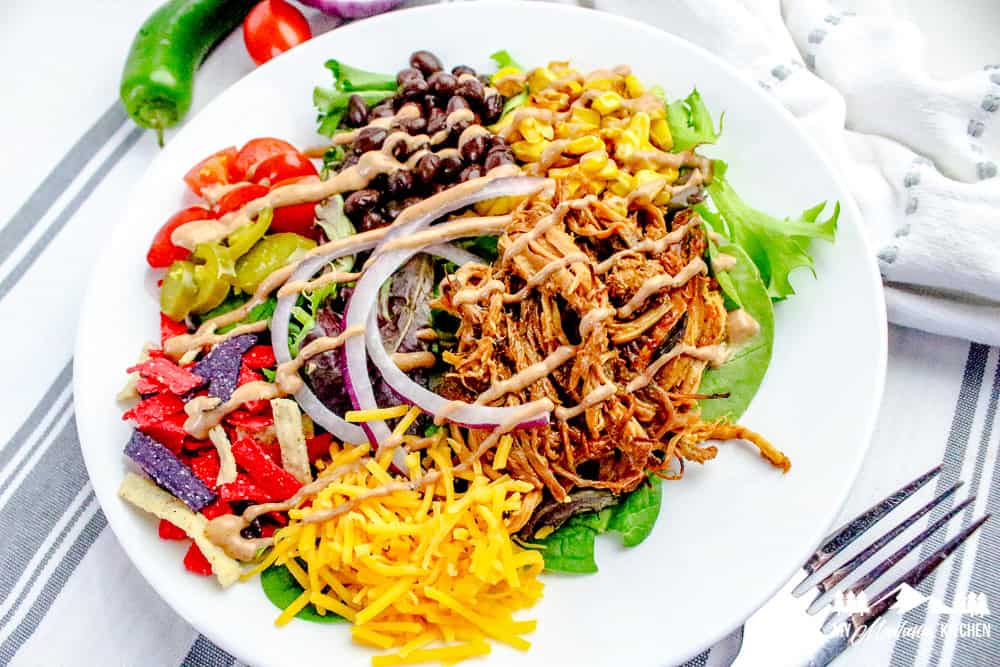 Finished pulled pork salad with shredded pork, grilled corn, black beans, cheese, cherry tomatoes, jalapeños, tortilla strips, red onion, and BBQ ranch drizzle on a white plate with striped background.