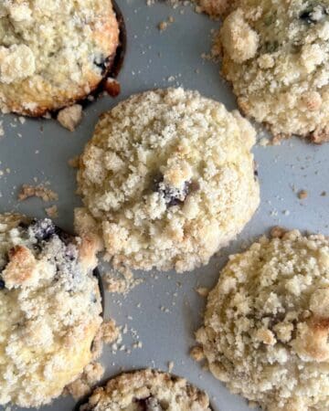 Close-up of freshly baked sourdough blueberry muffins in a muffin tin, topped with golden, buttery crumb topping.