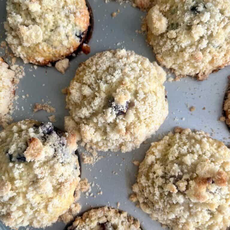 Close-up of freshly baked sourdough blueberry muffins in a muffin tin, topped with golden, buttery crumb topping.