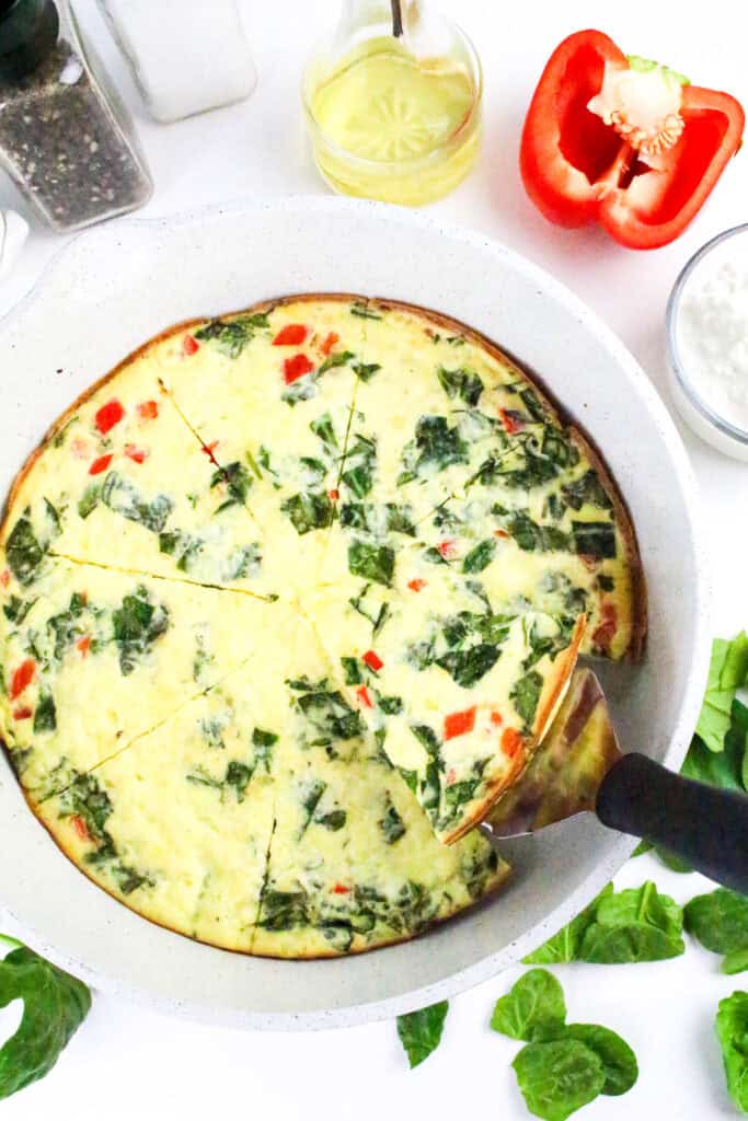 Baked cottage cheese frittata with spinach and red bell pepper in a white skillet, one slice being served.