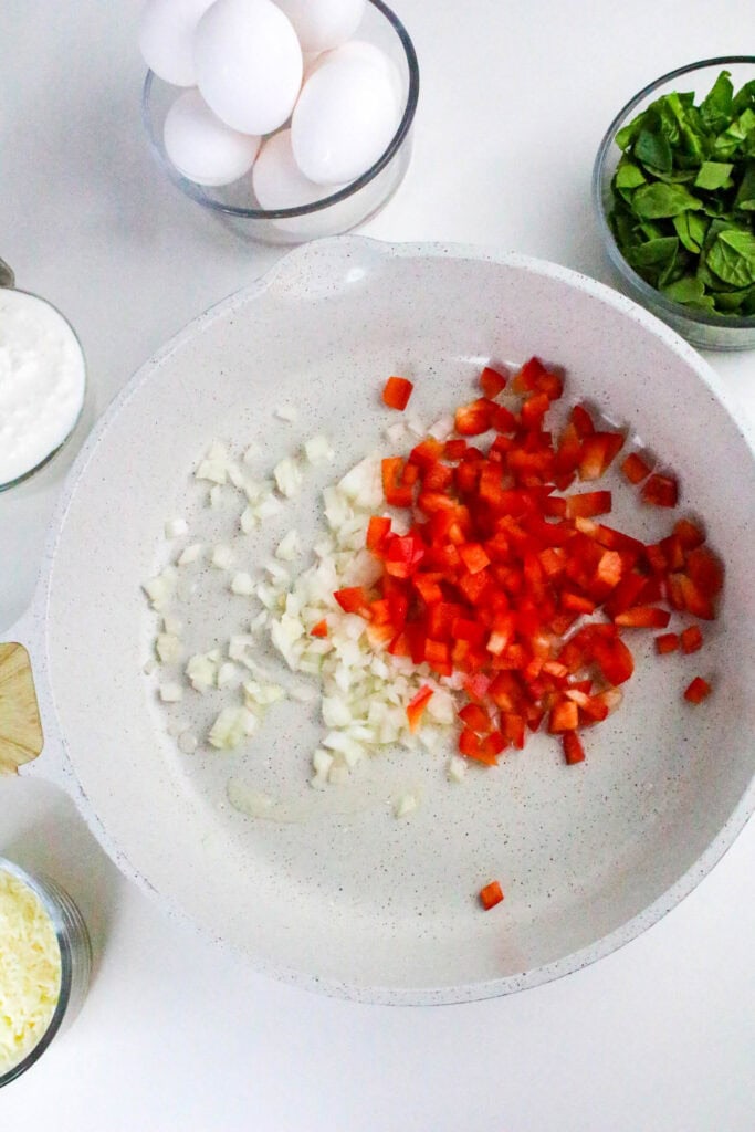 Chopped onions and red peppers cooking in skillet, surrounded by bowls of fresh eggs, spinach, and cottage cheese