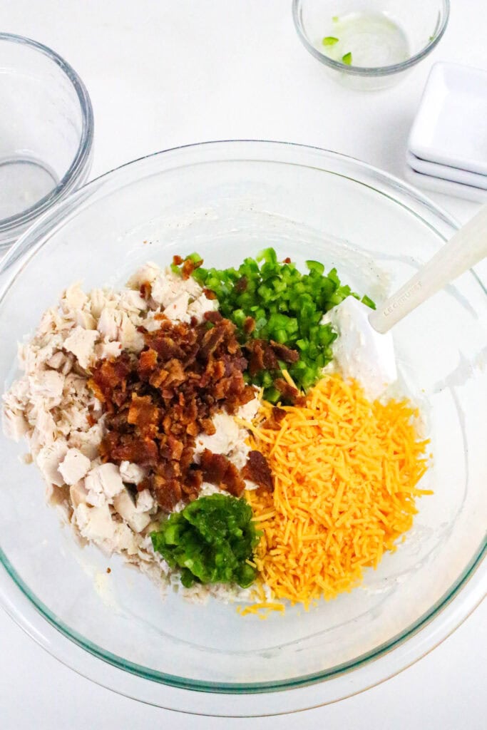 Glass bowl with layered ingredients for jalapeño popper chicken salad before mixing: diced chicken, bacon, cheddar, and jalapeños.