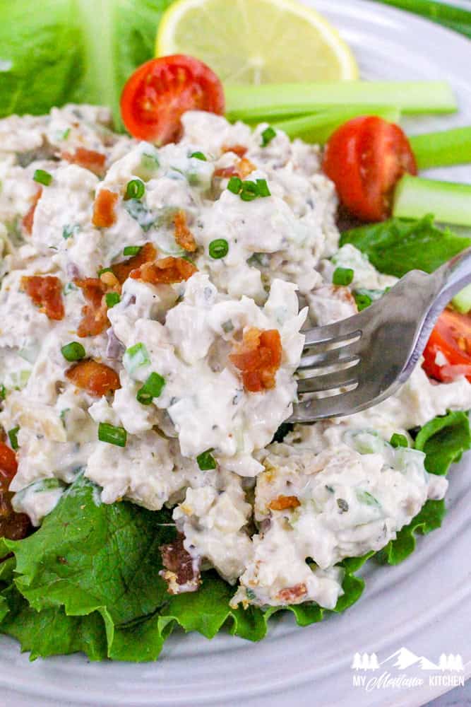 Fork lifting a scoop of creamy bacon ranch chicken salad with lettuce, cherry tomatoes, and lemon slices in the background.