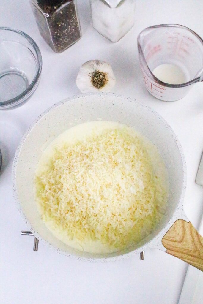 Grated parmesan cheese added to cream and garlic mixture in a skillet while making homemade low carb Alfredo sauce.