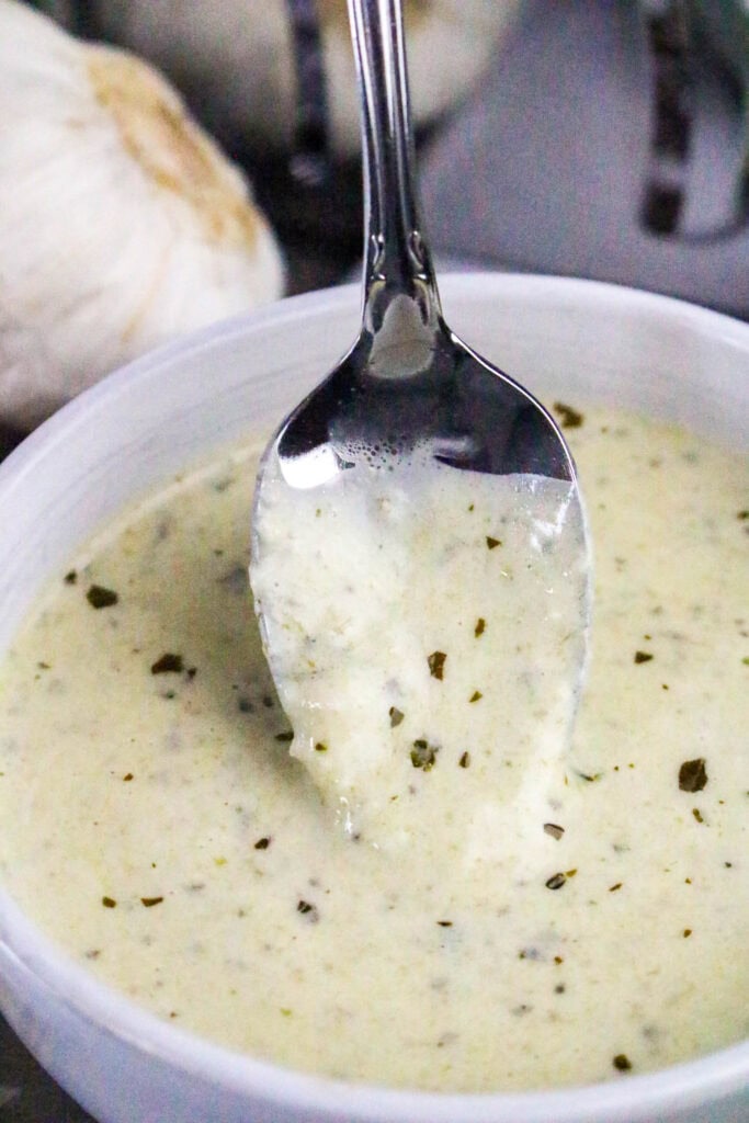 Close-up of spoon lifting creamy pesto Alfredo sauce from a bowl, showing smooth and velvety texture.