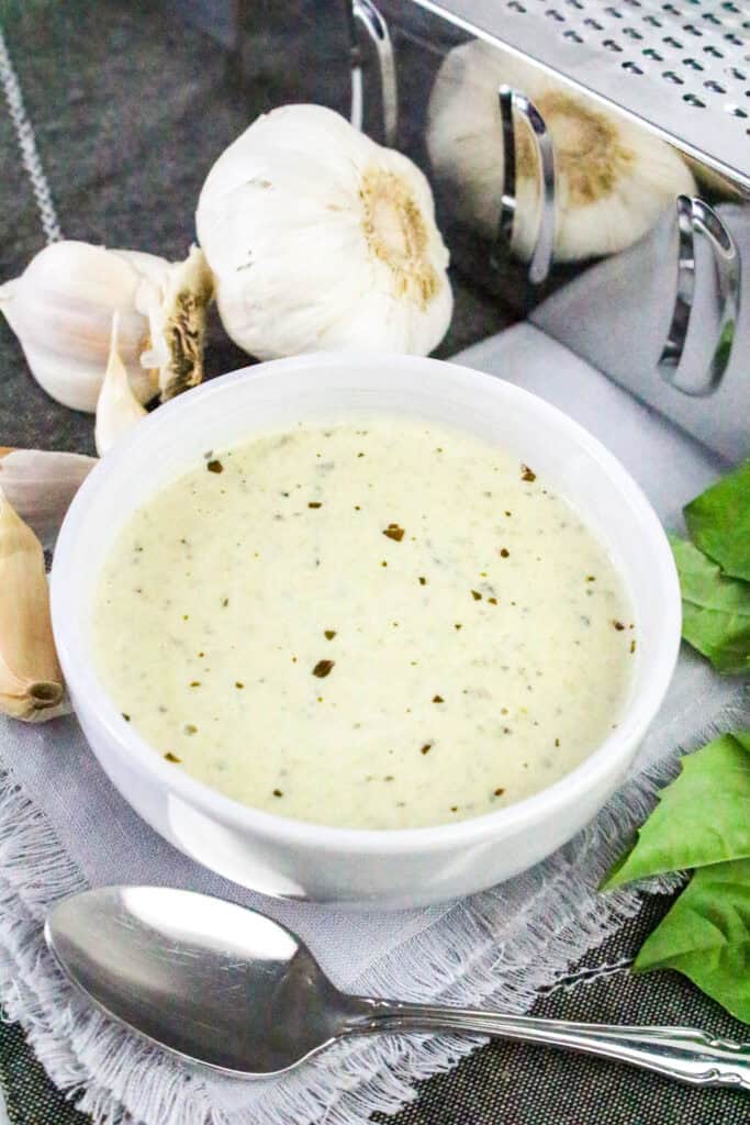 White bowl of creamy low carb pesto Alfredo sauce next to fresh garlic cloves and basil, with a spoon on a fringed napkin