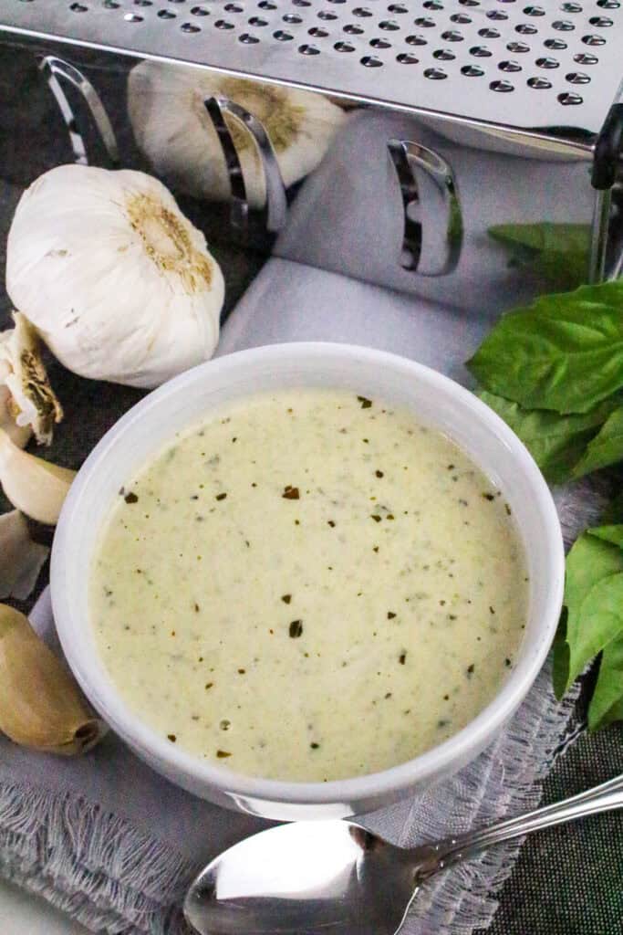 Overhead view of creamy basil pesto Alfredo sauce in a bowl, surrounded by fresh garlic, basil, and a cheese grater.