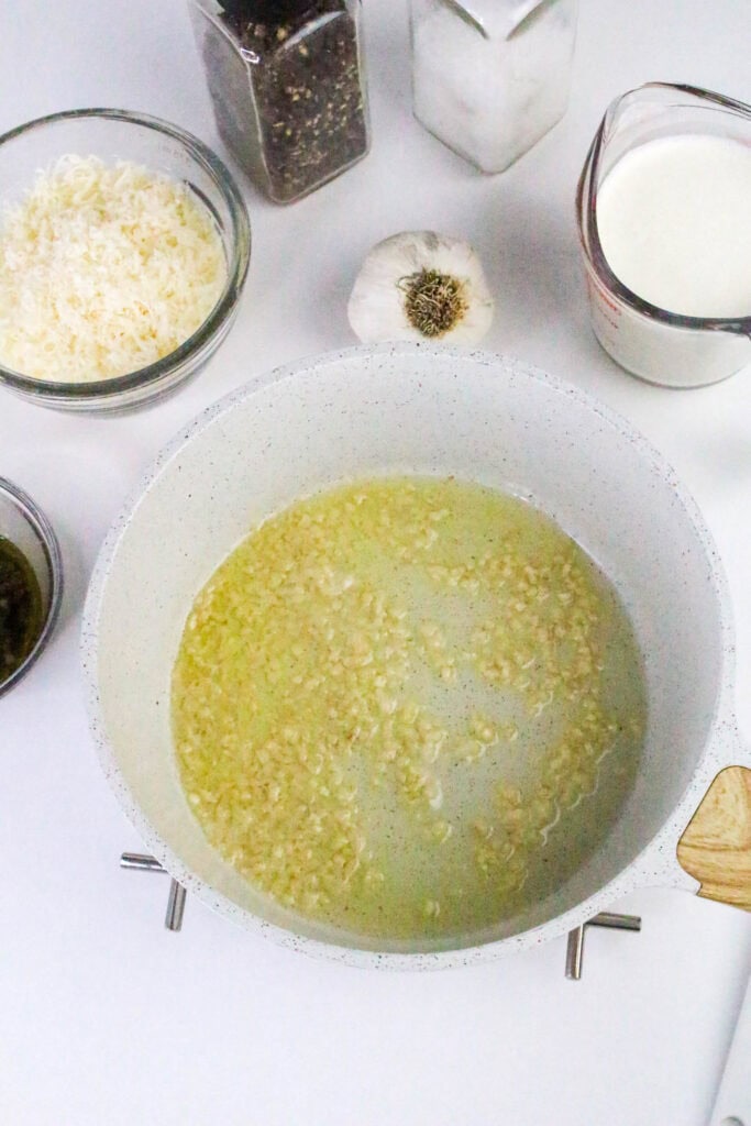 Garlic sizzling in melted butter in a white skillet, surrounded by ingredients for low carb creamy pesto Alfredo sauce.