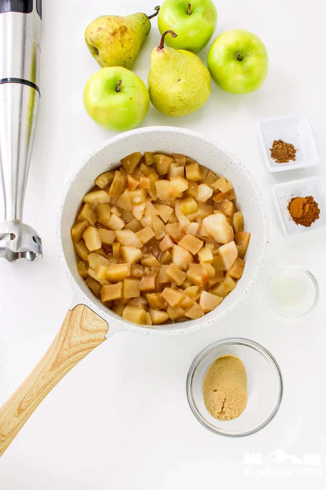 Cooked apples and pears in a pot with whole apples, pears, cinnamon, nutmeg, and sweetener nearby.