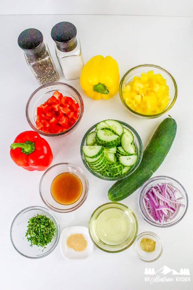 Flat lay of ingredients for bell pepper cucumber salad including cucumbers, bell peppers, onion, herbs, and dressing.