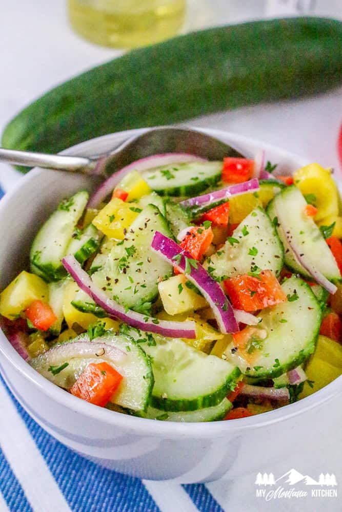 Fresh cucumber salad with red onion, red and yellow bell peppers, served in a white bowl with a spoon.