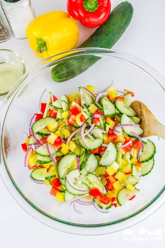 Overhead view of colorful bell pepper cucumber salad in a glass bowl with fresh ingredients nearby.