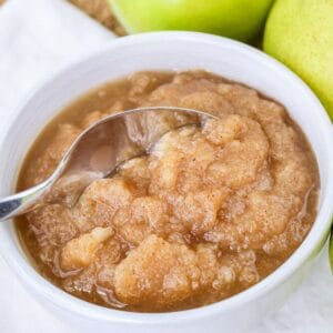 Close-up square image of a spoon scooping chunky apple pear sauce from a white bowl with green apples and pears beside it.