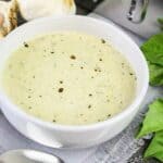 Bowl of thick and creamy basil pesto Alfredo sauce surrounded by garlic cloves and fresh basil leaves.