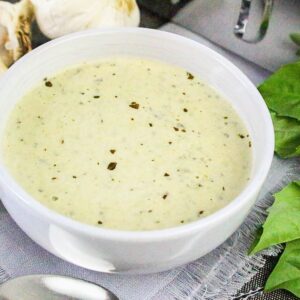 Bowl of thick and creamy basil pesto Alfredo sauce surrounded by garlic cloves and fresh basil leaves.