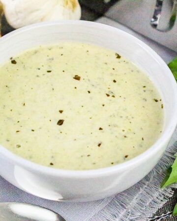Bowl of thick and creamy basil pesto Alfredo sauce surrounded by garlic cloves and fresh basil leaves.