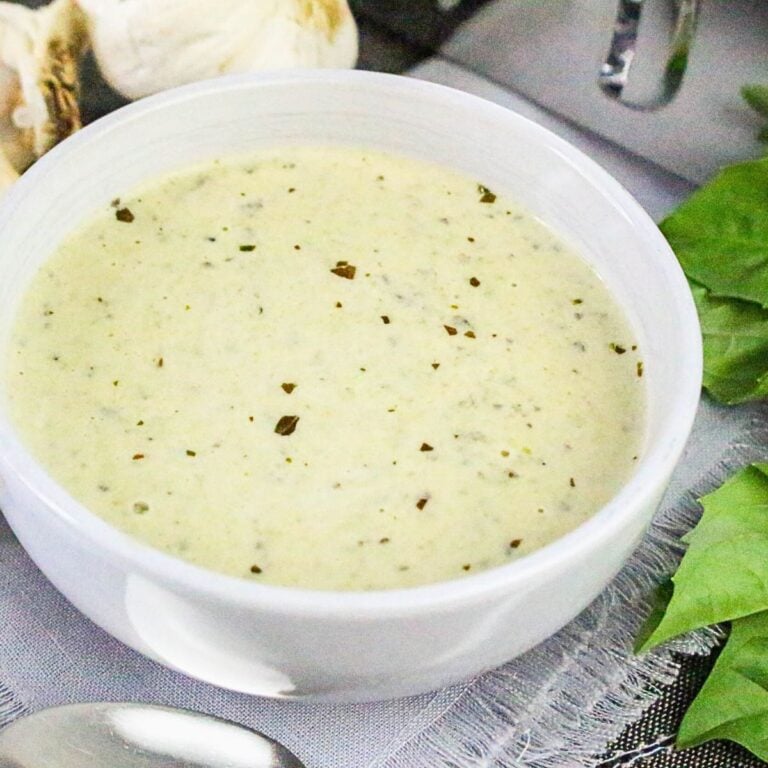 Bowl of thick and creamy basil pesto Alfredo sauce surrounded by garlic cloves and fresh basil leaves.