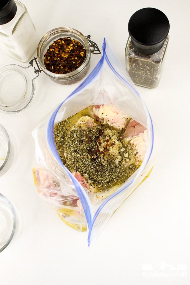 Italian herbs and seasonings being added to a zip-top bag of raw chicken thighs.