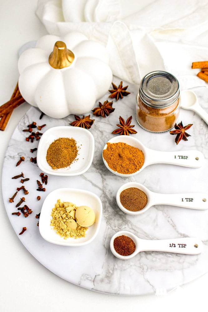 Flat lay of pumpkin pie spice ingredients in small bowls and spoons, styled with mini pumpkins, cloves, and cinnamon