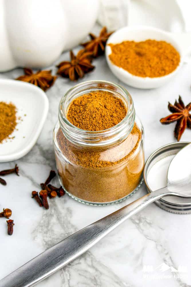 Glass jar of homemade pumpkin pie spice on marble counter, surrounded by cloves, star anise, and cinnamon sticks