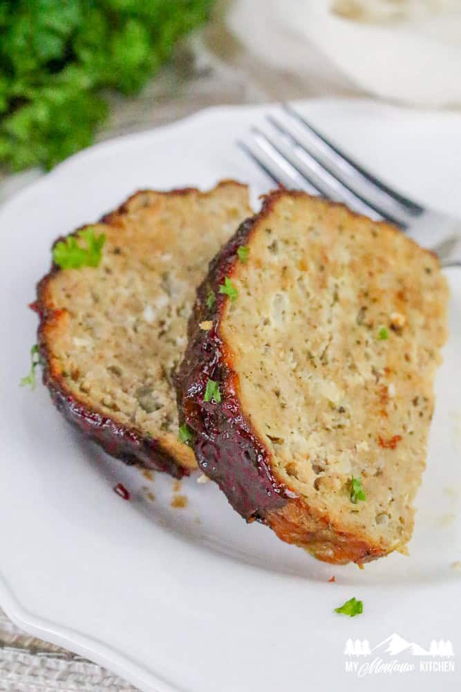 Two cleanly cut slices of cooked turkey meatloaf on a plate with a silver fork and fresh herbs in the background