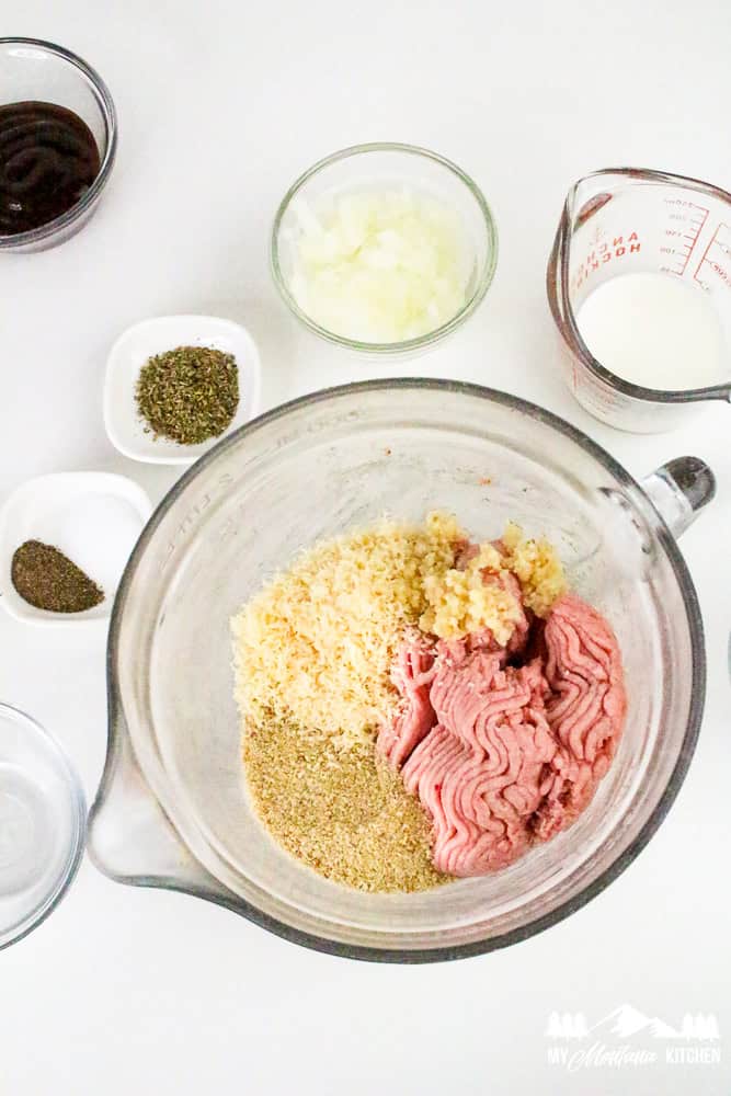 Top-down view of a glass mixing bowl with ground turkey, Parmesan cheese, garlic, breadcrumbs, and other classic meatloaf ingredients